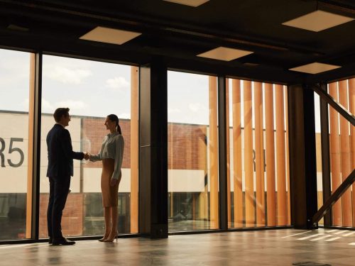 Wide Angle View Of Real Estate Agent Shaking Hands With Client While Standing In Empty Office Building Interior Lit By Sunlight, Copy Space
